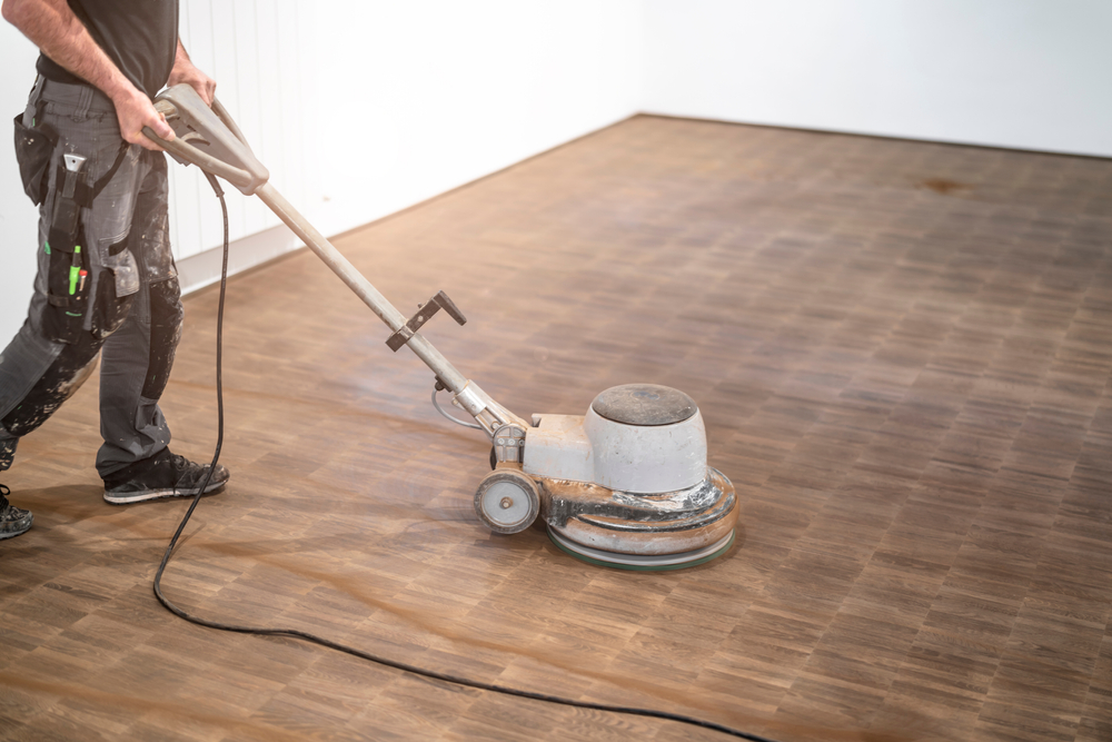 A person using a professional floor sander to restore a wooden floor, creating a visible difference between the treated and untreated areas.