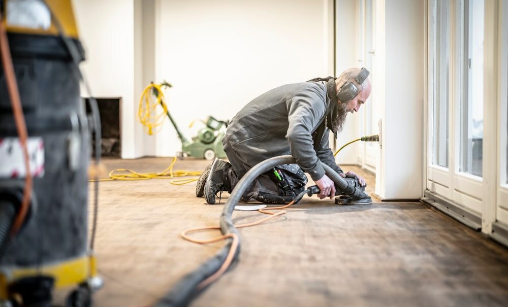 A professional Mr Sander® is operating the Festool Rotex 150mm sander on a herringbone floor. The sander, with dimensions of 150mm and a powerful 2kW effect, is connected to a dust extraction system with a HEPA filter for cleanliness. 