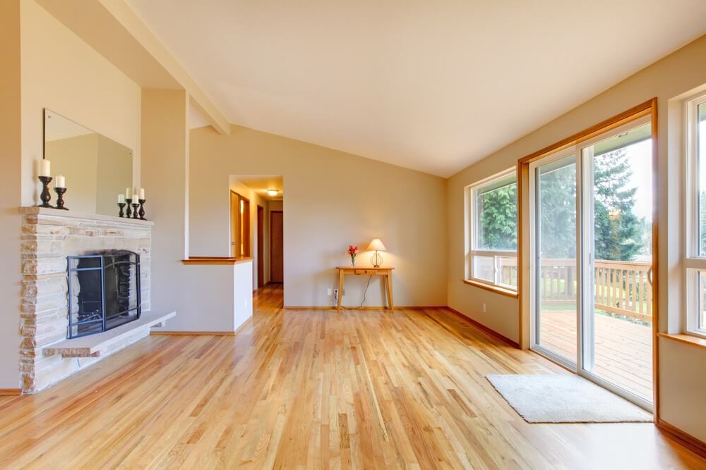 A spacious living room with large windows and a sliding glass door leading to a deck. The room features a newly sanded and refinished oak hardwood floor by Mr Sander® in Mitcham, UK. The light-colored oak flooring adds warmth and elegance to the space, complemented by a stone fireplace with candles and a small wooden table with a lamp in the corner.