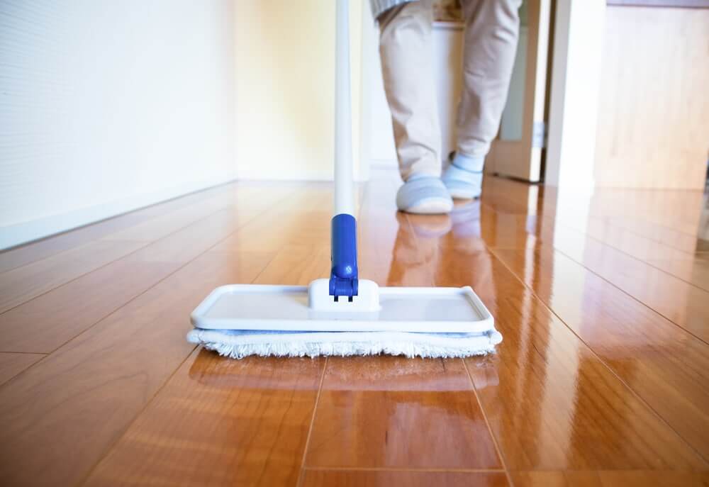 A person using a microfiber mop to clean a shiny, freshly refinished wooden floor. The polished surface reflects light, showcasing the excellent refinishing work done by Mr Sander®.