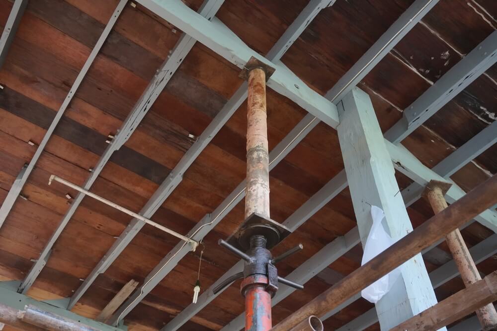A view of a wooden ceiling structure undergoing renovation, featuring dark-stained wooden planks supported by light blue beams. A metal jack post is used to stabilize the framework, indicating ongoing repair and restoration work.