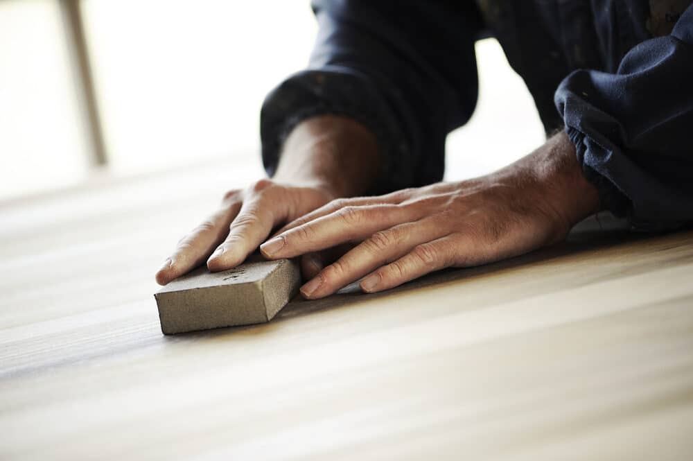 A person’s hands sanding a wooden floor with a block of sandpaper