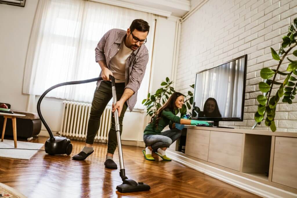 A man vacuums a beautifully restored parquet floor while a woman dusts the TV stand in a bright living room. The image highlights the importance of regular maintenance and cleaning to keep parquet floors looking pristine and elegant.