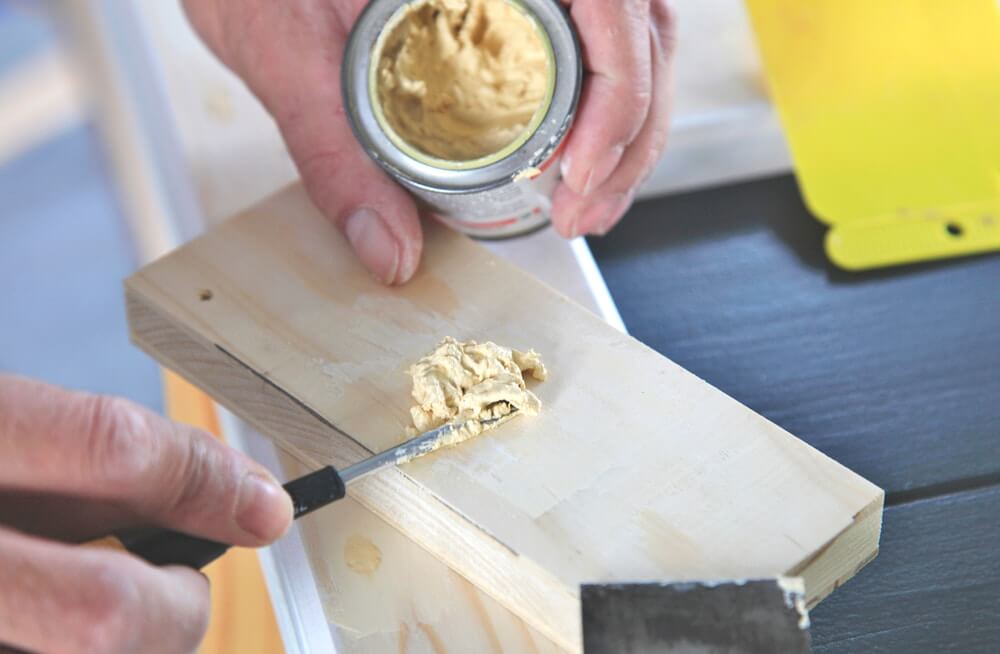 A close-up image of a person applying wood filler to a piece of wood using a small spatula. The filler is being scooped from a small container, demonstrating the preparation stage before sanding and refinishing a wooden floor.