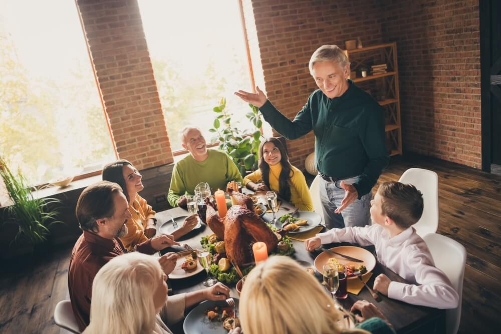 A family gathered around a dining table in a loft-style room with exposed brick walls and large windows. They are enjoying a meal, with a roast turkey as the centerpiece. The room features dark, polished hardwood floors that add warmth and contrast to the space.