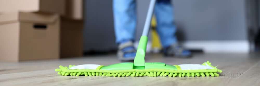 A close-up of a green microfiber mop being used on newly refinished oak hardwood floors. The floor displays a smooth and polished finish, achieved through professional sanding and refinishing by Mr Sander®. In the background, boxes and a person are visible, suggesting a final cleaning step after the refinishing process to remove any remaining dust or debris.