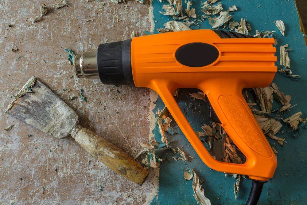 A heat gun and a paint scraper on a wooden surface with peeling paint.