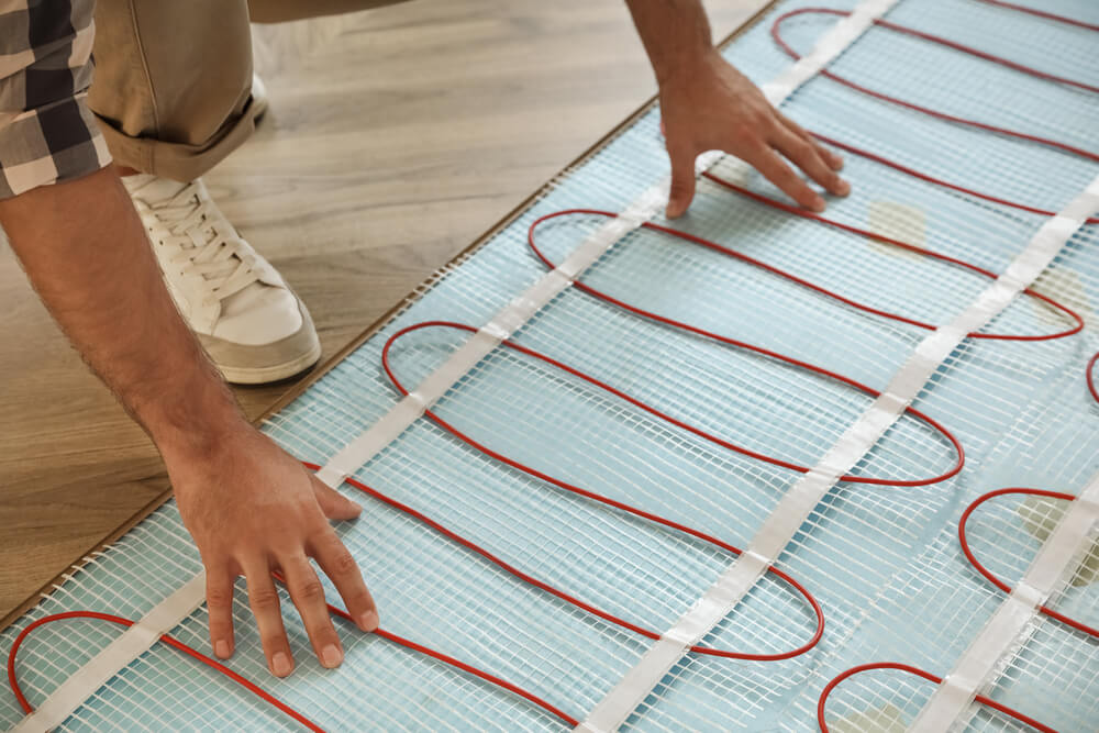A person installing underfloor heating in a home. They are carefully placing and securing red heating cables on blue mesh mats over a wooden subfloor, preparing for the installation of the final flooring layer.