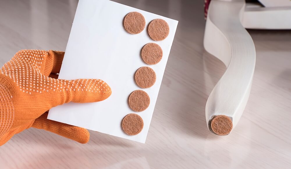 A person wearing an orange glove is holding a sheet of adhesive felt pads. One felt pad is already applied to the bottom of a chair leg, protecting a newly refinished wooden floor from scratches and damage. The floor has a smooth, glossy finish.