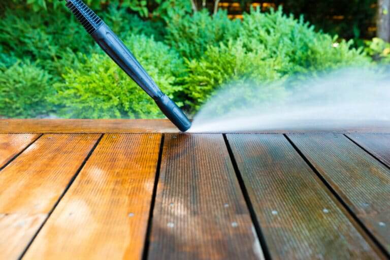 Close-up of a pressure washer nozzle cleaning a wooden deck, with a mist of water and a green garden in the background.
