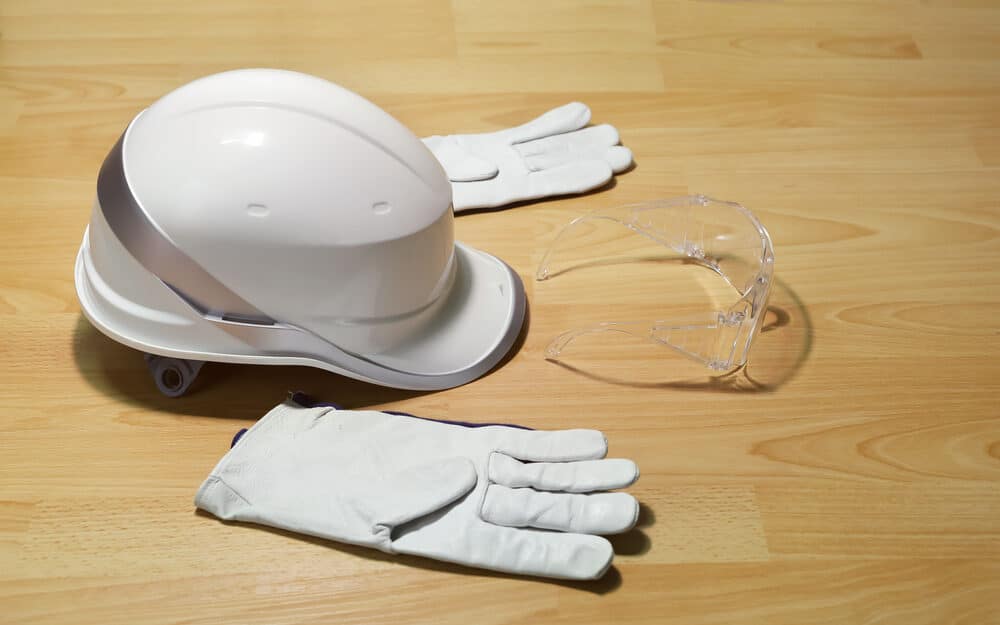 Personal protective equipment with a white hard hat, safety goggles, and gloves laid on a wooden floor.