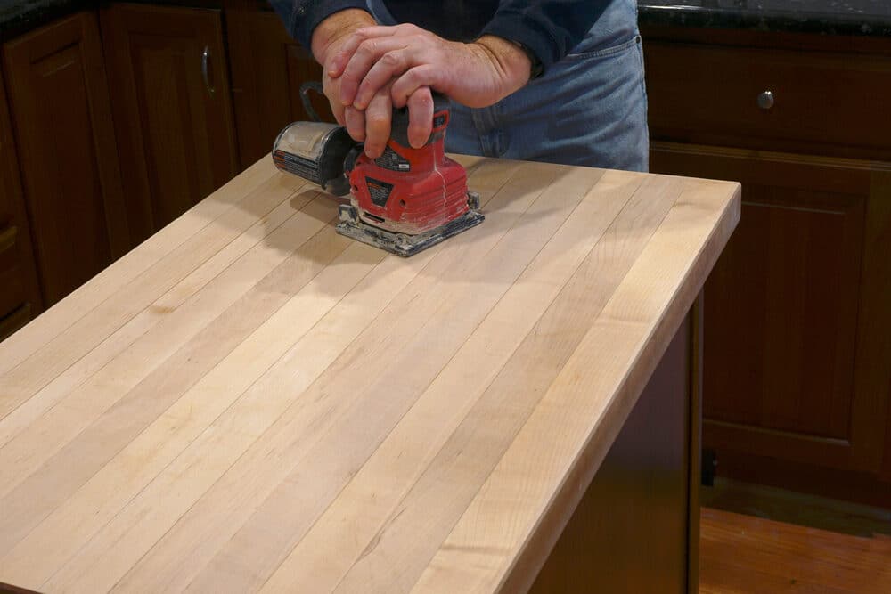 A person is sanding a wooden countertop with an orbital sander in a kitchen with wooden cabinets in the background.