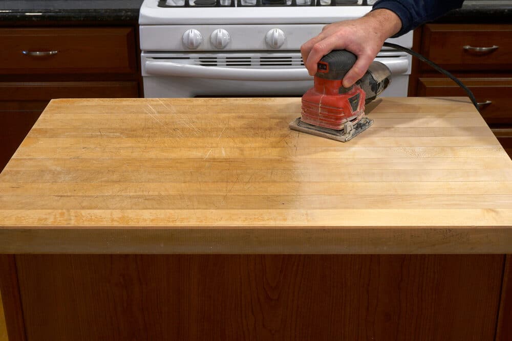 A person's hand holding an electric sander on a well-used wooden kitchen countertop with scratches and a stove in the background.