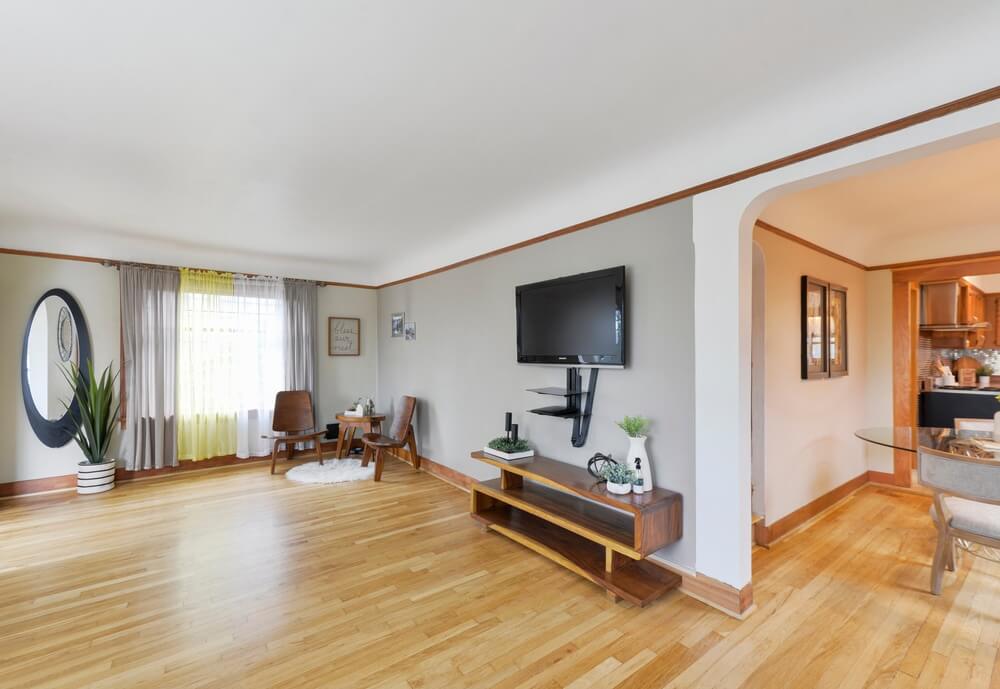 A bright living room with freshly sanded and refinished hardwood floors. The room features a mounted flat-screen TV, a wooden bench with plants, two chairs by the window, and a round mirror on the wall. The floor sanding and refinishing was done by "Sanding Wood Floors" in Southgate, UK.