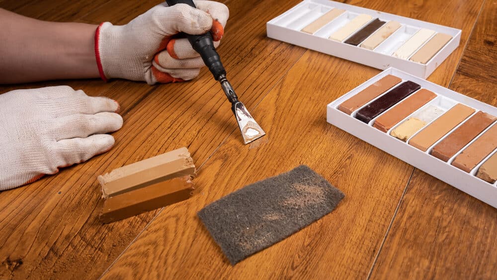 Hands in gloves using a putty knife with wood filler and sandpaper on a wooden surface.