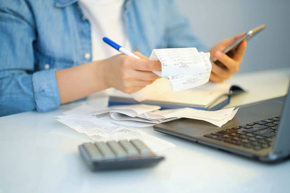 Person managing finances with receipts, a calculator, and a smartphone, alongside a laptop on a desk.