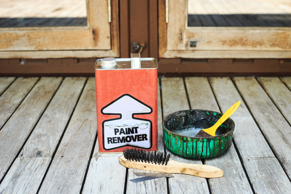 Paint remover can, a used brush, and an open tin with a yellow spatula on a wooden deck.