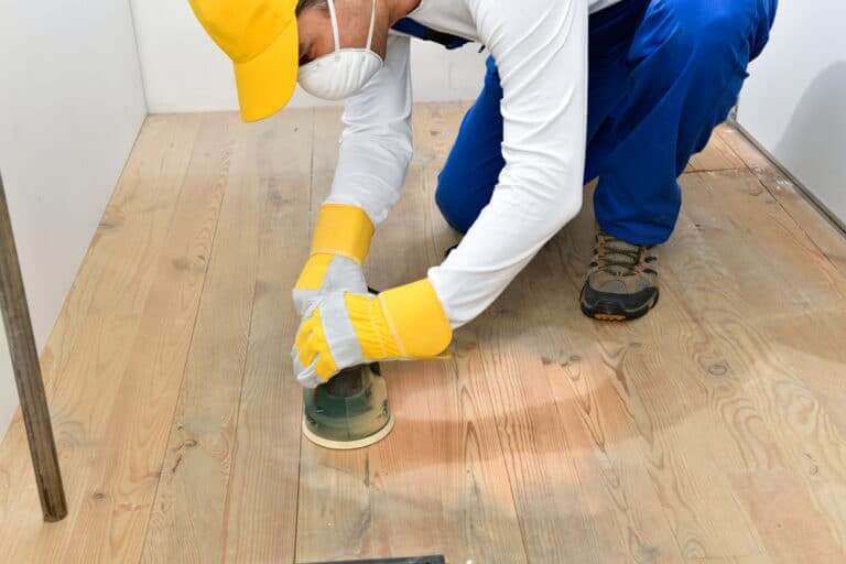 A worker wearing protective gear using a handheld sander on a natural wood floor.