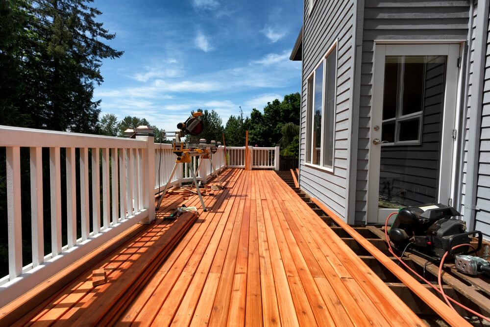 A partially completed outdoor deck made of freshly sanded and refinished cedar wood. The deck features white railings and is adjacent to a gray house. Tools and equipment are visible, indicating ongoing construction work.