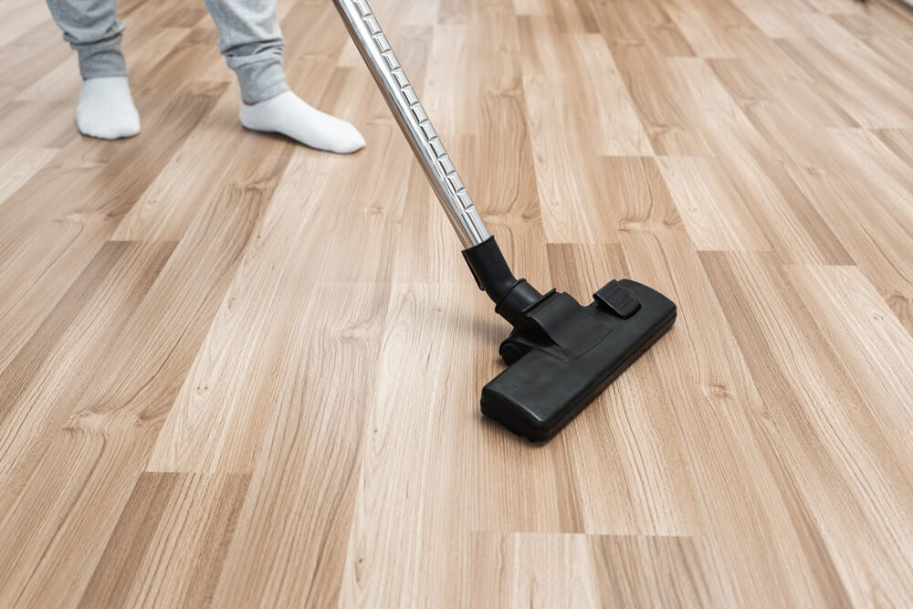 A person using a vacuum cleaner on a newly sanded wooden floor, ensuring the surface is clean and ready for refinishing. The person's feet are visible, wearing socks and standing on the smooth, light-colored wood.