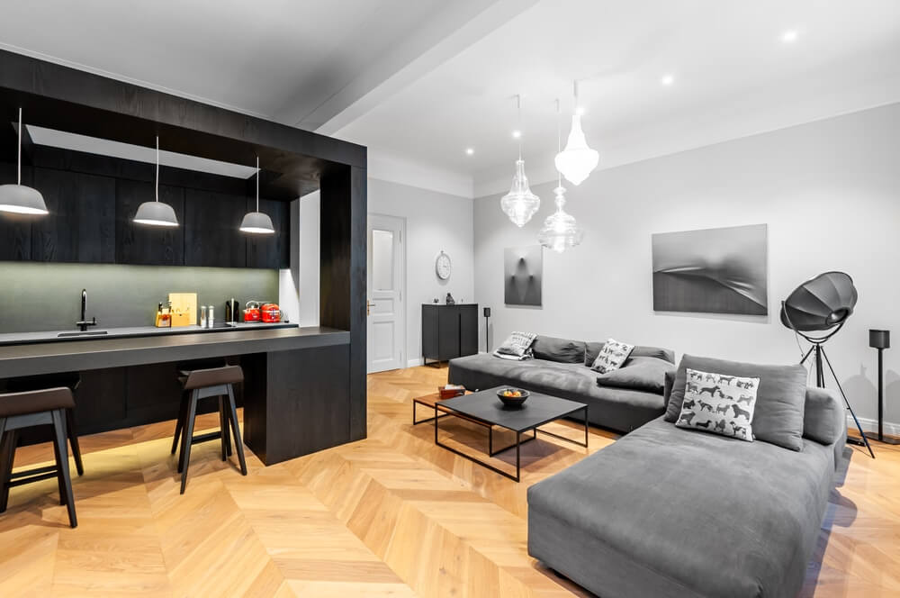 A modern open-plan living room and kitchen with a newly sanded and refinished herringbone parquet floor by Mr Sander®. The room features a sleek black kitchen island with bar stools, grey sofas, contemporary lighting fixtures, and minimalist decor. The floor's smooth and polished finish enhances the room's sophisticated look.