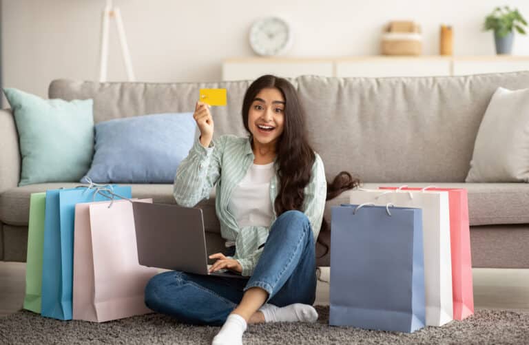 A happy young woman sitting on the floor of her living room in Haringey, London, surrounded by colorful shopping bags, holding a credit card and using a laptop, enjoying her online shopping experience.