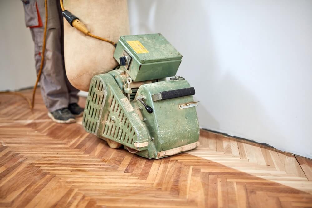 A Mr Sander® technician sanding a parquet floor in a Chadwell Heath living room using a LAGLER HUMMEL® machine. Features include powerful LED lighting, high powered aggressive sanding, extremely low dust emissions, quick belt changes, 30+ years lifespan, and easy maintenance. Technical specs: 230 V / 50 CPS / 2.2 kW motor, 200 mm drum width, 2400 rpm drum speed, 200 x 750 mm belt dimensions, 79 kg weight.