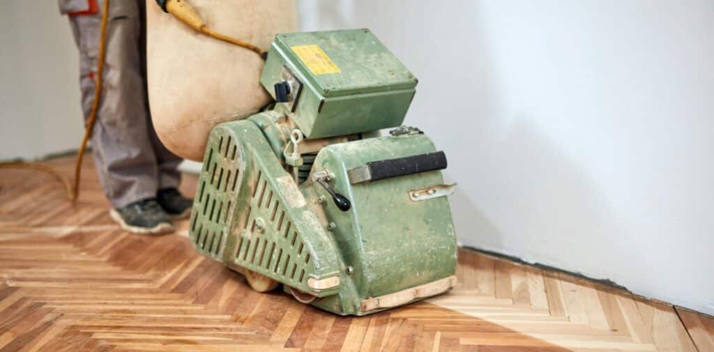 A man from Mr Sander® sands parquet flooring in a living room in East Acton, UK, using a Lagler HUMMEL. This powerful machine features a 300mm sanding width, ideal for large areas in public buildings. Technical specs include a three-phase AC motor with no-voltage release and thermal overload protection, operating at 400V/50CPS/4.2kW. It supports both star and delta voltage connections, with a sanding drum width of 300mm rotating at 2400rpm. The sanding belt measures 300 x 800mm, and the machine weighs 87kg.