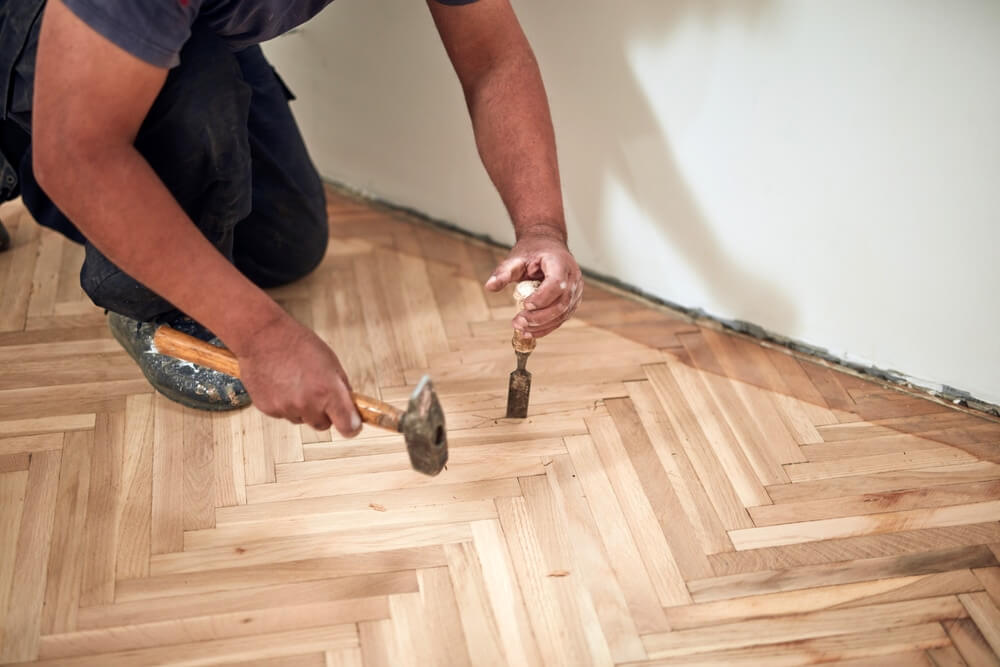 A professional from Mr Sander® using a hammer and chisel to repair a herringbone parquet floor during a sanding and refinishing project in a UK living room. The worker is kneeling, focusing on fitting and securing the wooden planks for a seamless finish.
