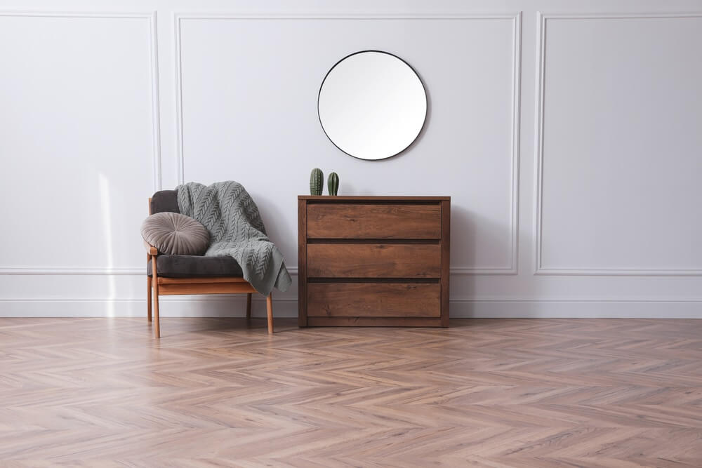 A stylish, minimalist room in Leytonstone, London, featuring newly sanded and refinished herringbone-patterned oak hardwood flooring by Mr Sander®. The space includes a wooden dresser, a cozy chair with a throw blanket and cushion, and a round mirror on the wall, showcasing the elegant flooring.