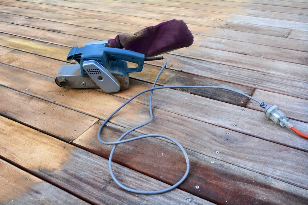 Electric hand planer left on a partially sanded wooden deck with power cord.