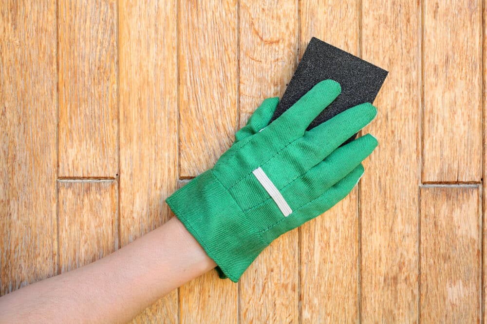 Hand wearing a green work glove holding a black sanding sponge against a wooden surface