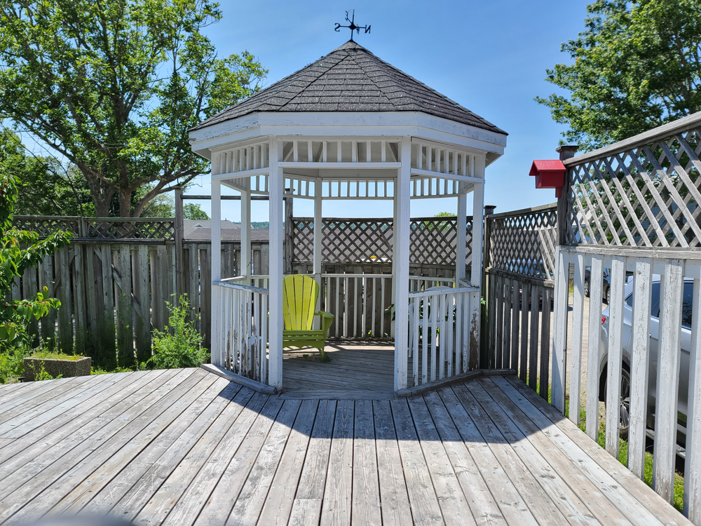 A wooden gazebo with a single green Adirondack chair on a weathered deck, enclosed by a white lattice fence, with trees and a clear blue sky in the background.