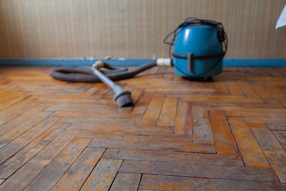 An old parquet floor is being vacuumed and cleaned before restoration. A blue vacuum cleaner and hose are seen on the floor, which shows signs of wear, dirt, and discoloration. This image illustrates the initial cleaning process necessary for effective parquet flooring restoration.