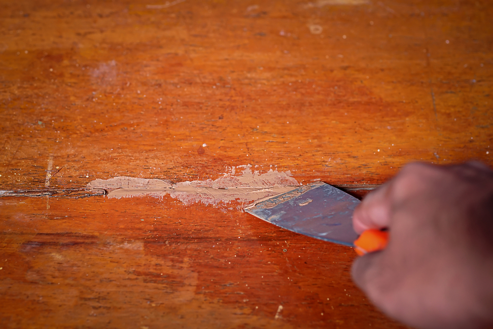 A hand holding a scraper removing paint from a wooden surface.