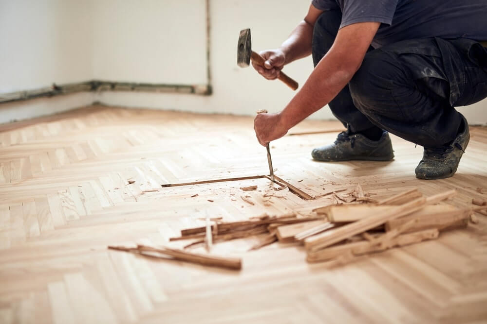 A professional from Mr Sander® is using a hammer and chisel to carefully remove damaged sections of parquet flooring as part of the restoration process. The image shows wood shavings and removed parquet pieces, emphasizing the detailed work involved in parquet flooring restoration.