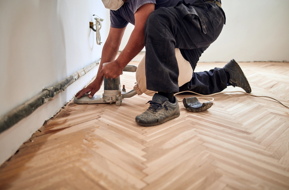 A professional working on sanding a parquet floor with a hand-held edge sander near a wall.