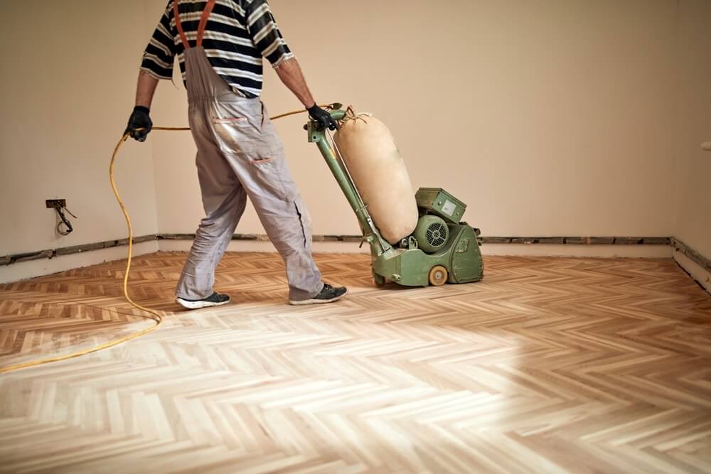 A man from Mr Sander® is sanding a parquet floor in a living room in West Brompton, UK, using the Lagler HUMMEL. This machine features a powerful LED lighting system, high powered aggressive sanding, and extremely low dust emissions. It has a single-phase AC motor with 230 V / 50 CPS / 2.2 kW & thermal overload protection, a sanding drum width of 200 mm, drum speed of 2400 rpm, sanding belt dimensions of 200 x 750 mm, and a machine weight of 79 kg.