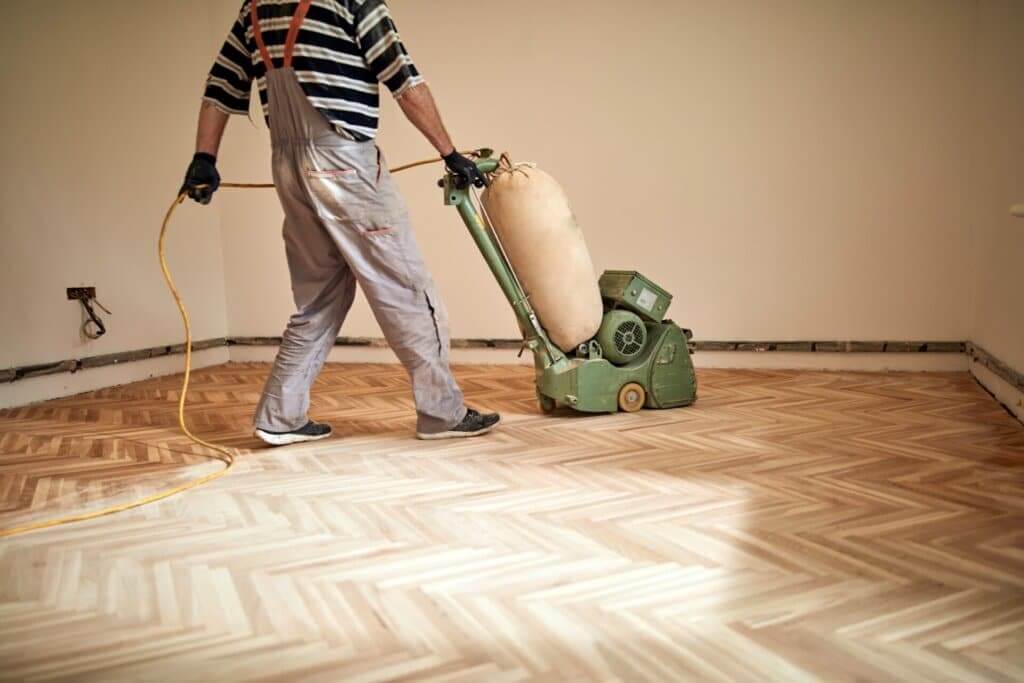 A worker using a professional sanding machine to restore a herringbone-patterned wooden floor in a room under renovation.
