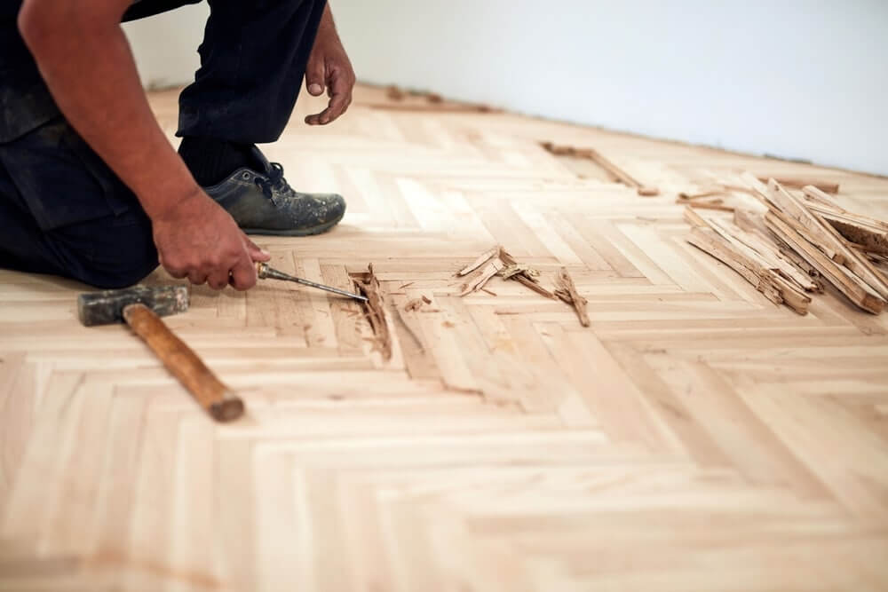 A professional from Mr Sander® kneeling and using a chisel to remove damaged sections of a herringbone parquet floor during a sanding and refinishing project in a UK living room. The floor is partially disassembled, with wood pieces and a hammer visible.