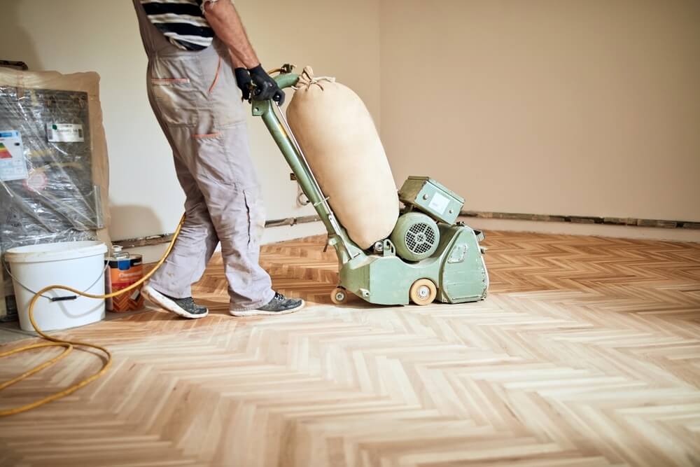 A professional from Mr Sander® sanding a floor in a living room in the UK using a Lagler HUMMEL® floor sanding machine. The machine features a powerful LED lighting system, aggressive sanding capabilities, low dust emissions, quick belt changes, and adjustable pressure. It's ideal for both large and small areas, ensuring optimal sanding quality.