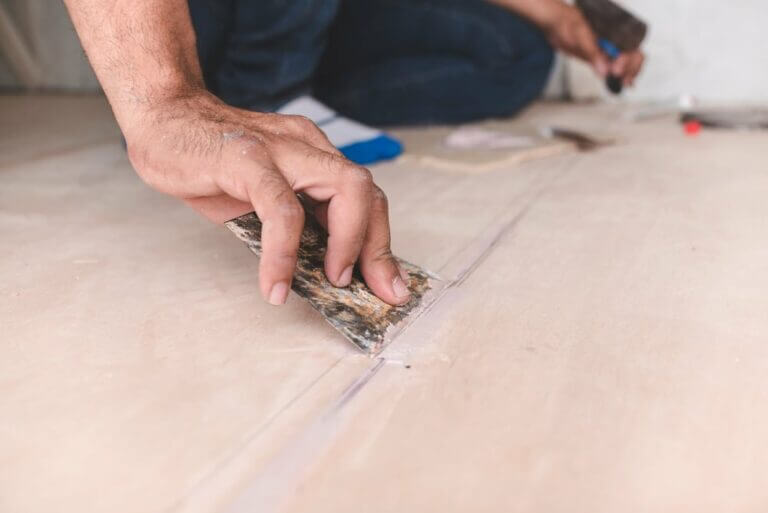 Close-up of a hand using a putty knife to spread wood filler into a gap on a wooden floor. Tools and materials for floor restoration are visible in the background.