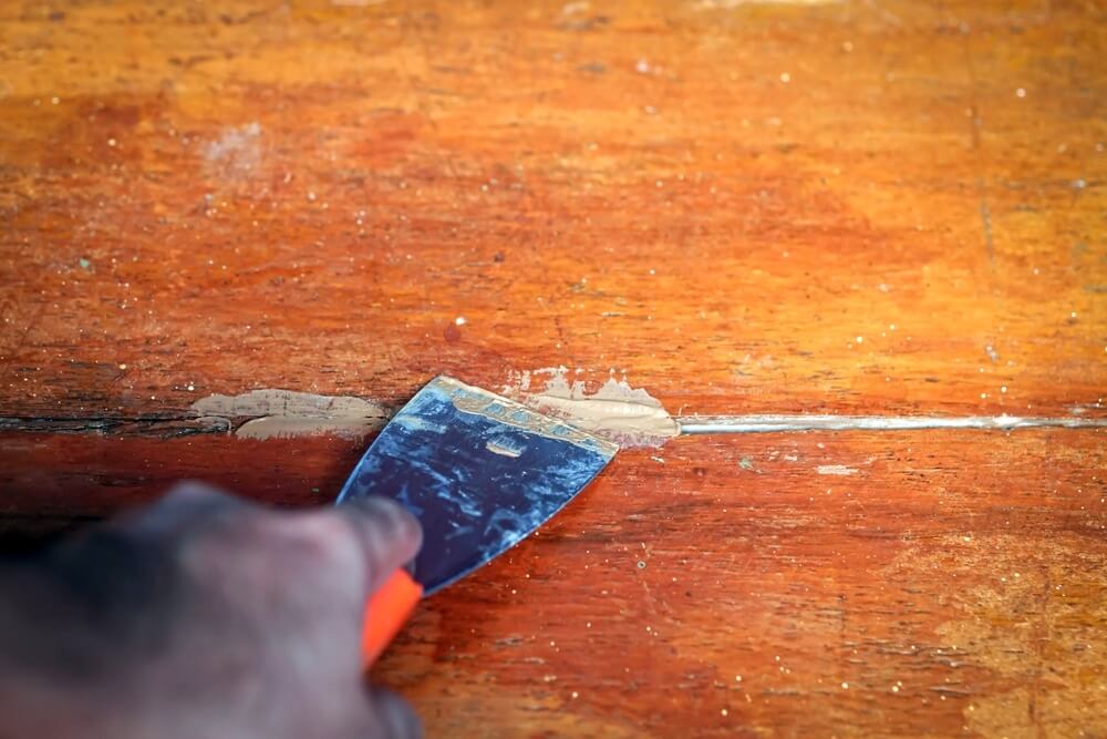 A hand using a putty knife to fill a gap in a wooden floor with wood filler during the floor sanding and refinishing process by Mr Sander®.