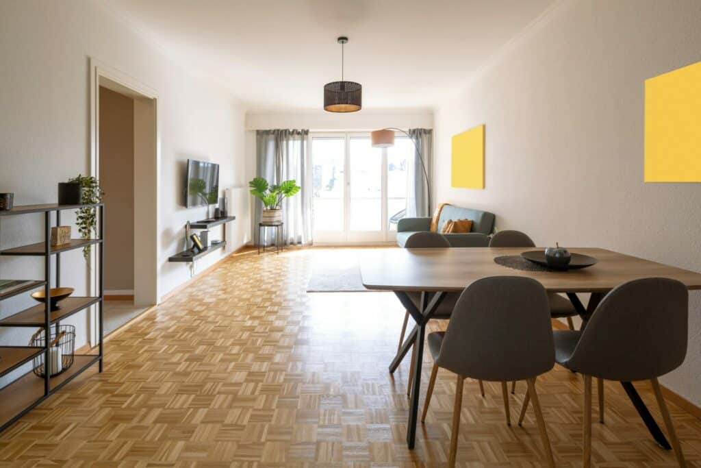 A spacious and bright living room with a newly refinished parquet floor that glows in natural light. The room features a modern dining area with a wooden table and grey chairs, a cozy sitting area with a green couch, and a flat-screen TV mounted on the wall. A large window with curtains lets in ample sunlight, highlighting the clean and polished floor.