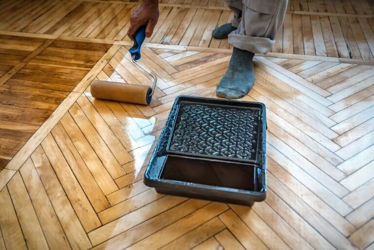 A Mr Sander® technician refinishing a parquet floor using a roller and tray. The partially sanded floor shows a clear difference between the refinished and unfinished sections. The technician is wearing socks to protect the floor while applying the finish.