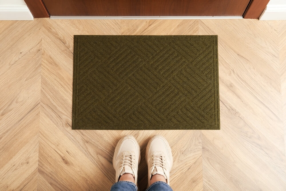 A pair of beige shoes standing on a freshly restored parquet floor next to a dark green doormat at the entrance. The image emphasizes the importance of using doormats to protect parquet flooring from dirt and damage, contributing to its maintenance.