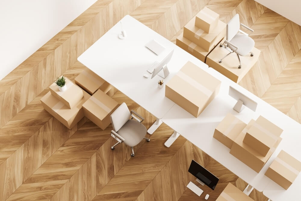  A modern office space with a chevron-pattern parquet wood floor, beautifully sanded and refinished to showcase its natural light wood tones. The image features a white desk with office chairs and cardboard boxes placed on top and around the workspace, suggesting a moving or setup phase.