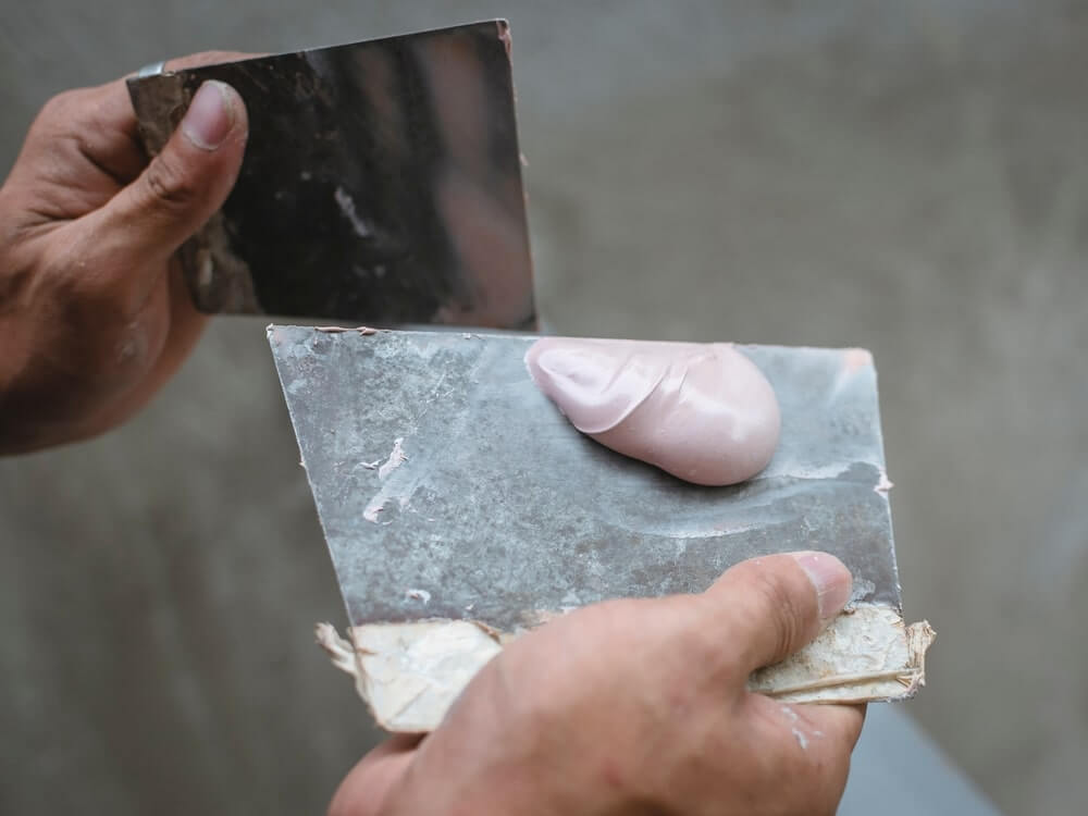 Close-up of hands holding two metal spatulas with a dollop of wood filler ready to be applied, demonstrating the preparation stage before floor sanding and refinishing.
