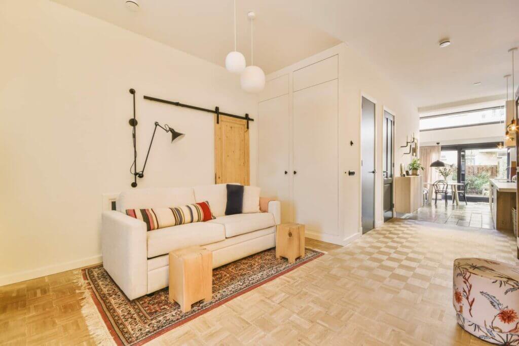 A modern living room with a light-colored parquet floor, recently sanded and refinished by Mr Sander® in Milton Keynes, UK. The room features a white sofa with colorful pillows, a decorative rug, and contemporary furnishings.