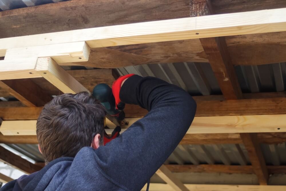 A worker using a power drill to fasten wooden beams during a floor restoration project, showcasing structural preparation.